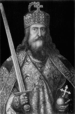 A portrait of Charlemagne. He has long hair and a beard, and wears richly
embroidered clothes and a jewelled and decorated crown. He holds an orb with a cross
in his left hand, and a raised sword in his right.